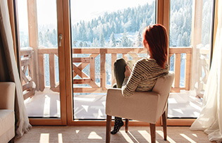Woman sitting in a comfortable chair drinking coffee and looking through window at snow covered mountain.